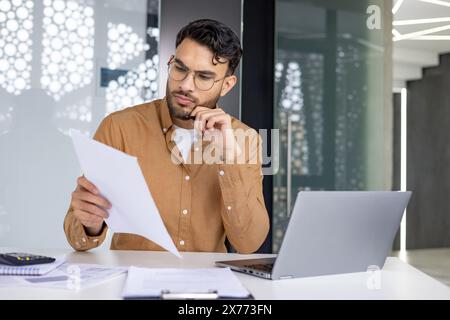 Young businessman concentrating on paperwork while working on a laptop. Professional setting in a modern office. Analyzing documents and making decisions. Stock Photo