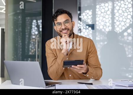Smiling man working at desk using a calculator in a modern office setting. Laptop and paperwork are visible on the desk. Stock Photo