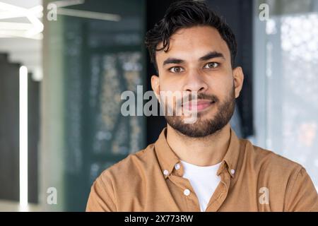 Confident young man with a beard smiling in a modern office environment, showcasing a professional and approachable appearance. Stock Photo