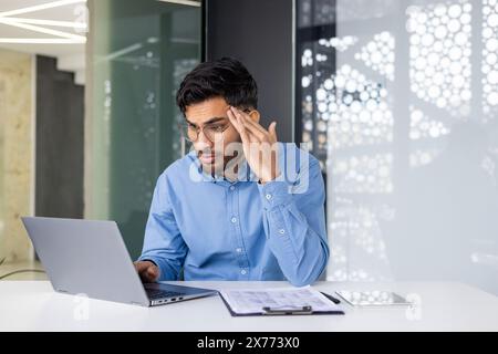 Young male professional in glasses and blue shirt looks stressed, working on a laptop in a modern office environment. Stock Photo