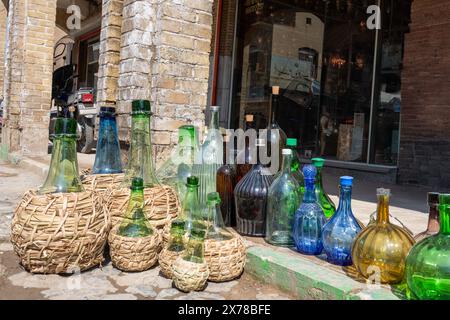 A variety of glass bottles, some in woven baskets, are displayed outside the store. Sunlight highlights their bright colors in Kashan, Iran. Stock Photo