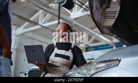 Experienced technician listens to customer in repair shop, using laptop to write down car modifications needed. Professional in garage with client, listening to her requests, zoom out shot Stock Photo