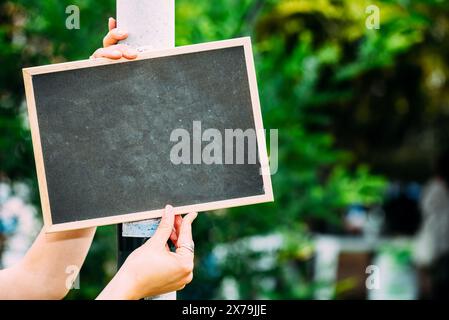 Someone is holding a blank chalkboard sign attached to a pole in a sunny park. The background is filled with lush green foliage Stock Photo