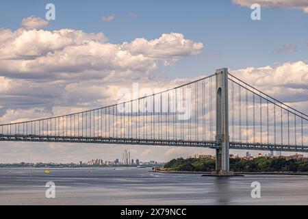 New York, NY, USA - August 1, 2023: Jersey City skyscrapers under Verrazzano-Narrows Bridge. Blue cloudscape above. Green Hamilton Fort park at shorel Stock Photo