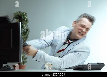 man in a light suit and striped tie, grimacing in frustration, wrestles with a computer monitor. The whiskey glass on his desk hints at his struggle w Stock Photo