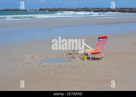 A single empty beach chairs sits on the sandy beach next to a sand castle covering in shells and a wave pool beside it, faces the ocean and rock jetty. Stock Photo