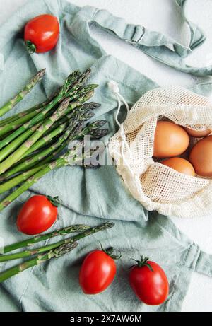 A bunch of fresh asparagus lies next to a reusable mesh bag filled with brown eggs, accented by ripe cherry tomatoes on a light-colored surface with a Stock Photo