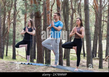 A group of three people doing yoga in the forest. A man and two women in a tree pose. Stock Photo