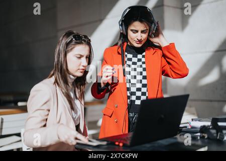 Focused female business professionals in smart casual attire collaborating on a project using a laptop and headphones on a sunny day outside. Stock Photo