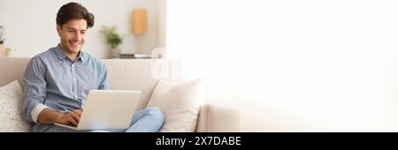 Happy young man sitting on couch, using laptop Stock Photo