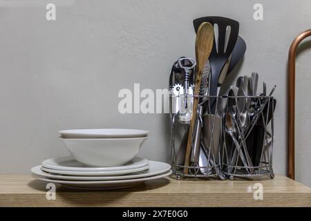 Selection of kitchen utensils on a shelf with plates and bowls stacked next to it against a textured wall as a background.  Copy space top left side. Stock Photo