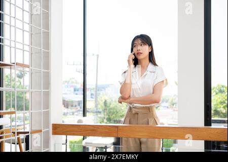 A confident, attractive millennial Asian businesswoman is having a serious phone conversation with someone in the company's hallway corridor. talking Stock Photo