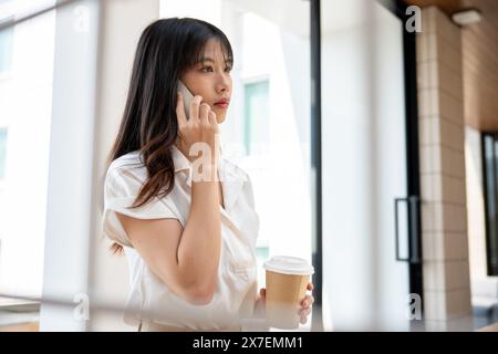 A confident, attractive millennial Asian businesswoman is having a serious phone conversation with someone in the company's hallway corridor during he Stock Photo