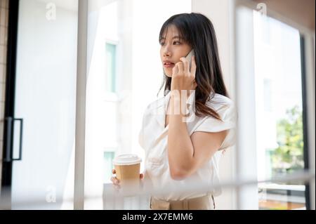 A confident, attractive millennial Asian businesswoman is having a serious phone conversation with someone in the company's hallway corridor during he Stock Photo