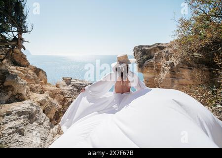 A woman in a white dress is standing on a rocky cliff overlooking the ocean. She is wearing a straw hat and she is enjoying the view. Stock Photo