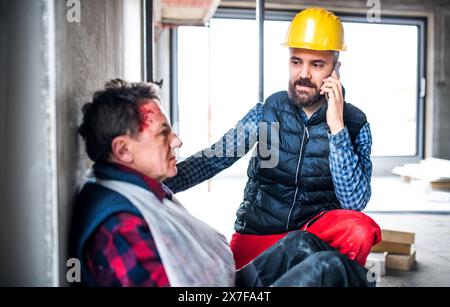 Colleague calling ambulance and giving first aid to injured worker, in pain after accident at construction site Concept of occupational safety and Stock Photo