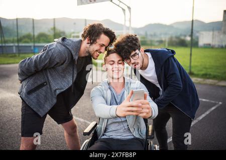 Young man in wheelchair spending free time outdoor with friends, taking selfie with smartphone. Male friendship. Stock Photo