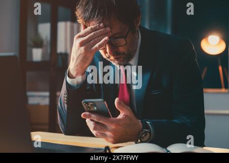 Disappointed businessman working in the office late at night, using cell phone and laptop computer, selective focus Stock Photo