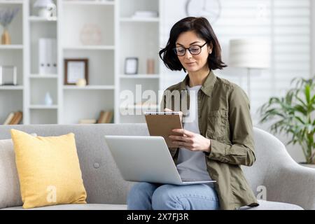 Young concentrated and serious woman sitting on the sofa at home and studying remotely, working on a laptop and making notes in a notebook. Stock Photo