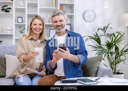 Happy mature couple using a smartphone for online banking while sitting on a sofa at home. They are handling finances together in a cozy living room. Stock Photo