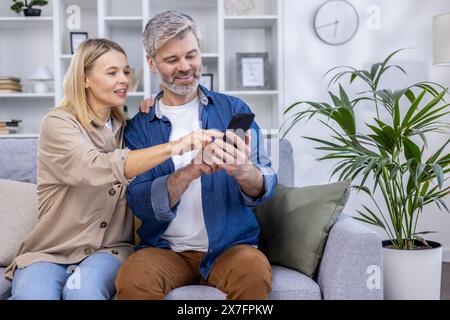 Smiling couple sitting on the couch at home, looking at a smartphone and enjoying quality time together. Stock Photo