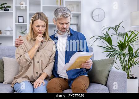 A concerned couple sitting on the couch, examining financial paperwork in their living room. They appear stressed while trying to understand the contents of the envelope. Stock Photo