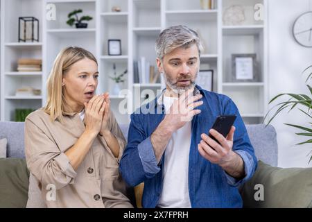 Middle-aged couple with shocked expressions, looking at a smartphone while sitting on the couch in a modern living room. Stock Photo