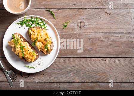 Scrambled Eggs Sandwiches and coffee for healthy keto breakfast. Whole grain toasts with scramble eggs and organic arugula microgreens on wooden table Stock Photo
