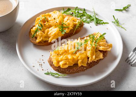 Scrambled Eggs Sandwiches and coffee for healthy keto breakfast. Whole grain toasts with scramble eggs and organic arugula microgreens on white table. Stock Photo