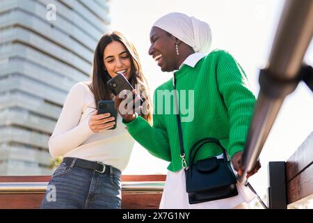 two young female friends laughing happy while having fun carefree looking at their mobile phones, concept of diversity and modern lifestyle Stock Photo