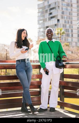 two young female friends laughing happy while having fun carefree using their mobile phones in a city park, concept of diversity and modern lifestyle, Stock Photo