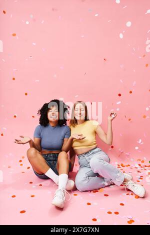 Two diverse women in cozy casual attire sitting on the ground surrounded by confetti. Stock Photo