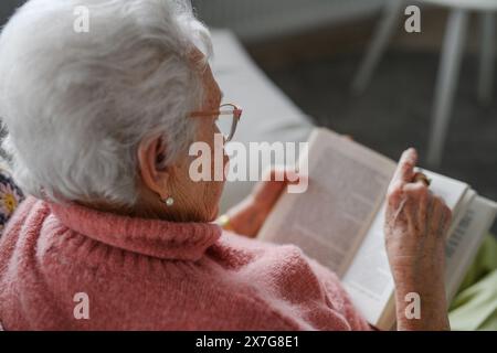Beautiful elderly woman reading book at home, having relaxing moment, hygge for senior. Grandmother with reading glasses and captivating book. Stock Photo