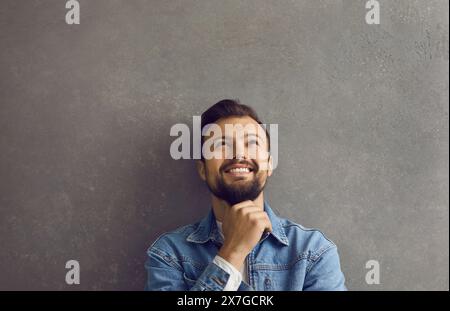 Happy young man leaning against gray wall, looking up, thinking, dreaming and smiling Stock Photo