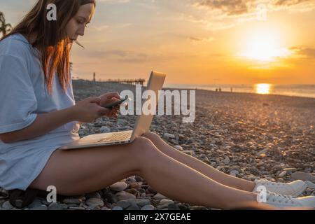 Digital nomad woman working with laptop and mobile phone on the beach by the sea during sunset. Female freelancer work online using smartphone and Stock Photo