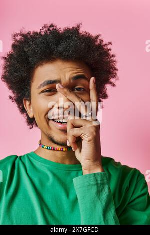 Cheerful African American man with curly hair in a green shirt, smiling and touching his face on a pink background. Stock Photo