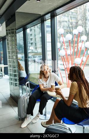 Two multicultural colleagues, diverse businesswomen, sitting at a table in a hotel, engaged in deep conversation in front of a window. Stock Photo