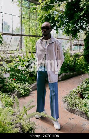 A stylish African American man stands confidently in front of a greenhouse filled with lush green plants. Stock Photo