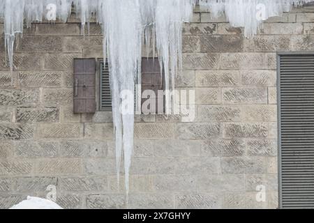 Large icicles hanging from the edge of a roof above a brick wall with two small windows and a vent. Stock Photo