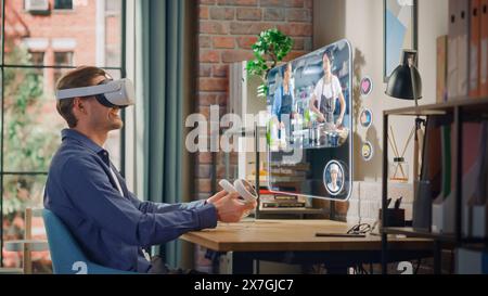 Handsome Young Man Sitting in Loft Living Room at Home, Using Virtual Reality Headset with Controllers to Check Social Media Streaming Application. Male is Watching a Cooking TV Show. Stock Photo