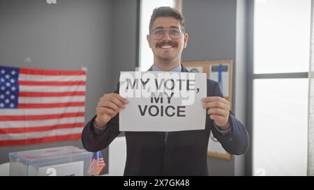 Smiling young hispanic man holding 'my vote, my voice' sign in a voting center with us flag. Stock Photo