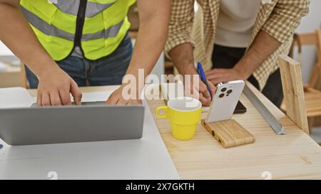 Two men collaboratively working in a carpentry workshop, using a laptop and taking measurements on a wooden table. Stock Photo