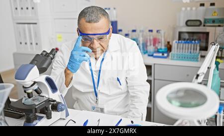 Hispanic man wearing lab coat and gloves analyzing samples with microscope in laboratory. Stock Photo