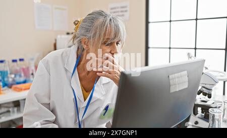 A pensive middle-aged woman in a lab coat analyzes data on a computer in a bright laboratory setting. Stock Photo