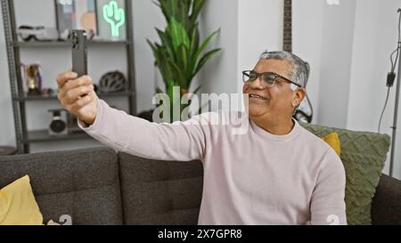 A smiling middle-aged man takes a selfie with his phone in a cozy, modern living room, showcasing an everyday moment. Stock Photo