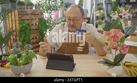 A middle-aged man with a moustache makes a payment during a tablet video call in a flower shop. Stock Photo
