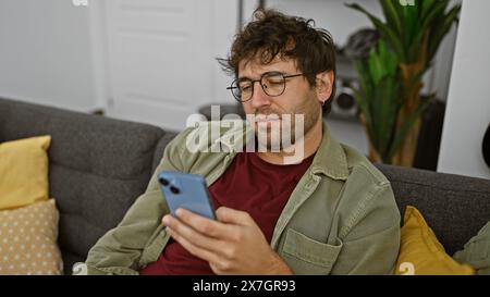 Hispanic man with beard and glasses using smartphone on couch indoors. Stock Photo