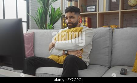 Handsome hispanic man with beard sitting on a gray sofa looking sad while watching tv in a cozy living room. Stock Photo