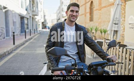 Handsome hispanic man in leather jacket smiling on motorcycle in sunny urban street scene. Stock Photo