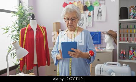 Senior woman tailor with glasses and measuring tape using tablet in colorful atelier. Stock Photo